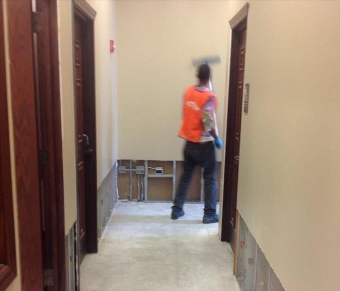 A SERVPRO crew member in an orange west standing at the end of a commercial hallway with flood cuts cleaning the walls.