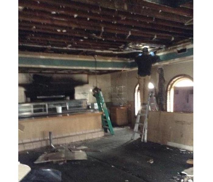 kitchen area of a restaurant with ceiling tiles removed, debris on floor, man on ladder